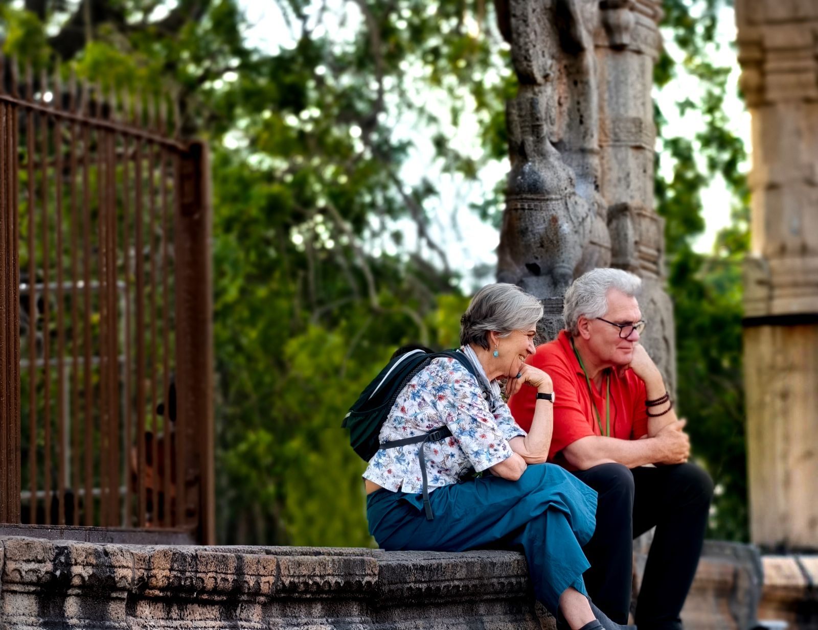 A senior couple resting on a stone wall at a historic site under shady trees