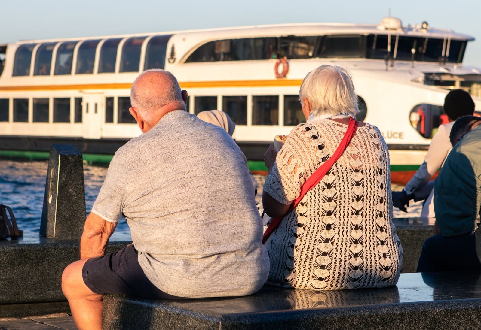 An older couple sitting on a waterfront wall watching a passenger boat go by, the easy pace of US river cruises for seniors